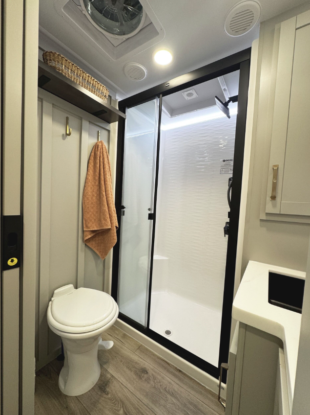 Modern RV bathroom featuring a corner shower with sliding glass doors and a black frame, a white toilet, light gray cabinetry, wood-look vinyl flooring, a ventilation fan above, and a hanging peach towel on gold hooks beneath a shelf with a wicker basket.