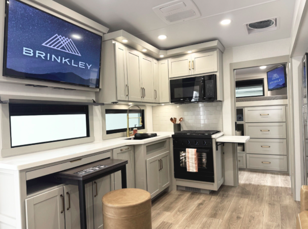 Interior of a luxury Brinkley RV featuring a modern kitchen with light gray cabinetry, white countertops, a black microwave and oven, under-cabinet lighting, a pull-out table, two large windows, and a mounted TV displaying the Brinkley RV logo.