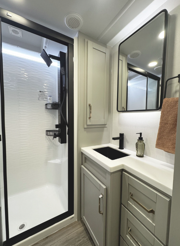 Stylish RV bathroom with a black-framed glass shower featuring a rainfall showerhead and built-in shelving, adjacent to a modern vanity with a black sink, matte black faucet, gold hardware, and a backlit mirror above. A peach towel hangs on a black towel ring next to a soap dispenser.
