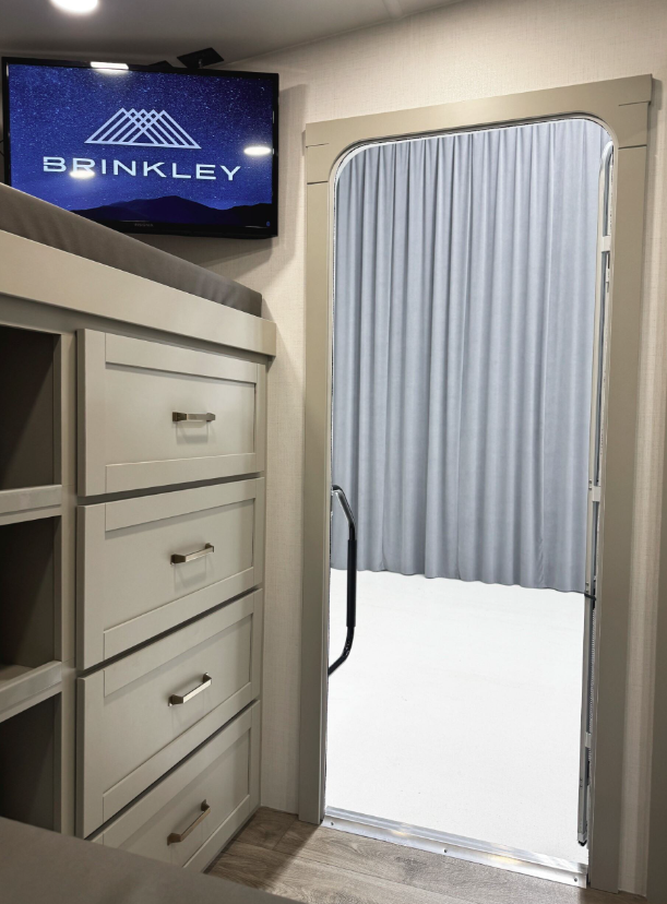 Interior view of a Brinkley RV showing a stylish dresser with four drawers and gold handles, a mounted flat-screen TV displaying the Brinkley logo, and an open door leading outside with a visible handrail and grey curtain backdrop.