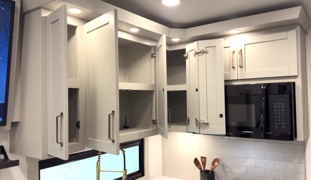 Upper kitchen cabinets in a Brinkley RV shown open to reveal generous storage space, with under-cabinet lighting, a black microwave, modern gold hardware, and a white subway tile backsplash creating a bright, functional cooking area.