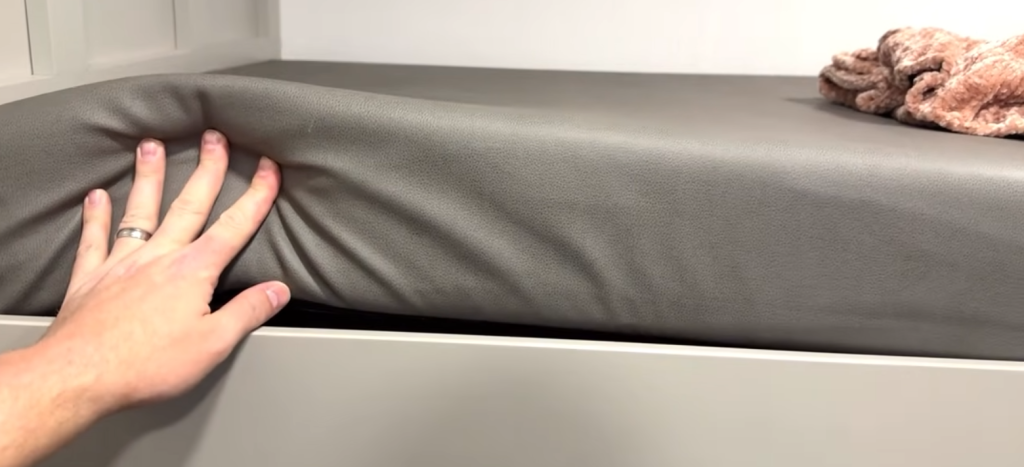 Close-up of a hand pressing down on a thick, plush bunk mattress in an RV, demonstrating its softness and support, with a cozy blanket resting at the corner.