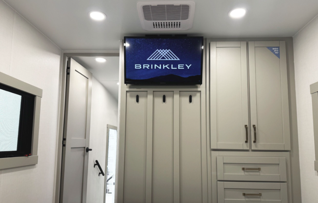 Interior of a Brinkley RV showcasing a mounted TV above a coat rack with storage hooks, overhead LED lighting, and adjacent built-in cabinetry for wardrobe storage.