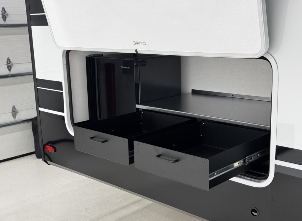 Close-up of an RV's exterior camp kitchen featuring a Furrion mini fridge and two pull-out metal storage drawers built into the side wall under a large hinged access door.