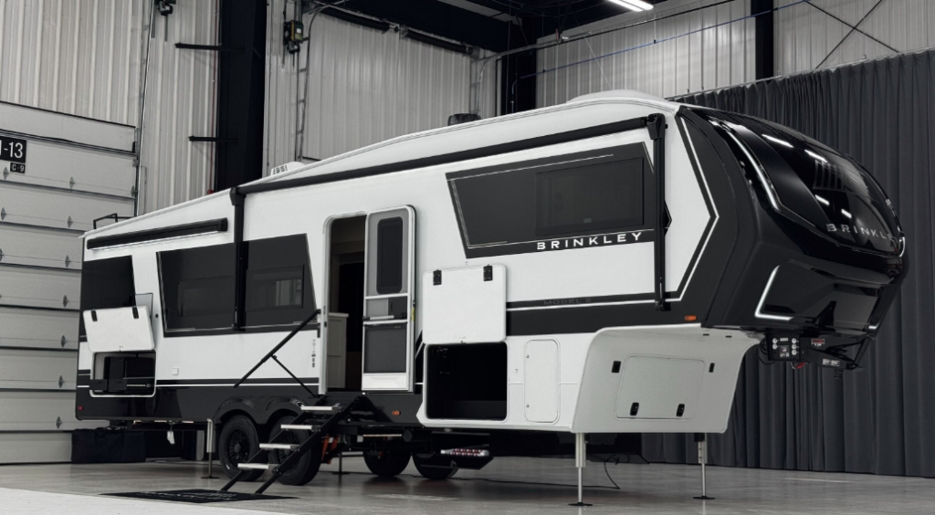 Brinkley Model Z fifth wheel RV displayed in an indoor showroom, featuring sleek black-and-white exterior design, multiple slide-outs, oversized tinted windows, and sturdy entry steps.