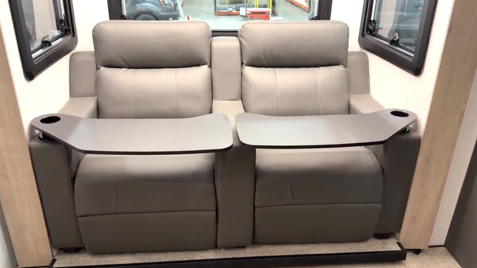 Dual reclining theater seats in an RV featuring light gray upholstery, individual cup holders, and detachable swivel tray tables, positioned in front of large windows for natural light and scenic views.