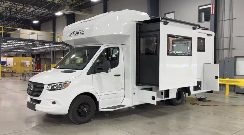Driver side view of a Grand Design Lineage 25TK Class C motorhome inside a factory, with slide-out extended, showcasing its white body on a Mercedes-Benz chassis, storage compartments open, and the "LINEAGE" branding above the cab.