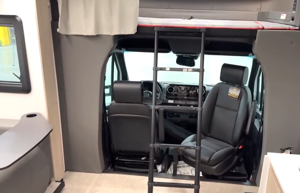 Interior view of a motorhome cab area with black leather driver and passenger seats, a central ladder leading to an overhead bunk with a privacy curtain, and part of a recliner and countertop visible on either side of the image.