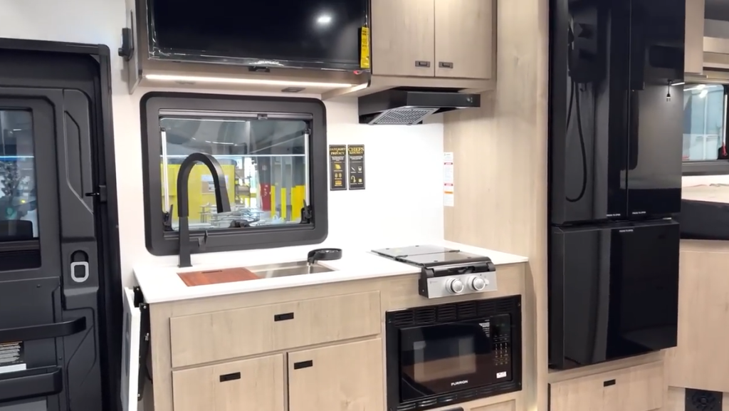 Close-up view of a Lineage RV kitchen showcasing a stainless steel sink with a black pull-down faucet, extended countertop with built-in cutting board, dual-burner stove, microwave, and open drawers and cabinets revealing additional storage space beneath the counter.