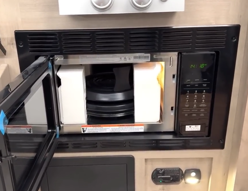 Close-up of an RV microwave installed below a stovetop, with the door open to reveal protective packaging inside, a turntable, and digital controls displaying the time 14:18; situated in a modern kitchenette setup with light wood cabinetry.