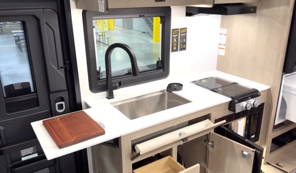 Interior kitchen of a Lineage RV featuring light wood cabinetry, a black high-arc faucet over a stainless steel sink, a dual-burner cooktop with range hood, a microwave, a large black double-door refrigerator, an overhead TV, and a window providing natural light.






