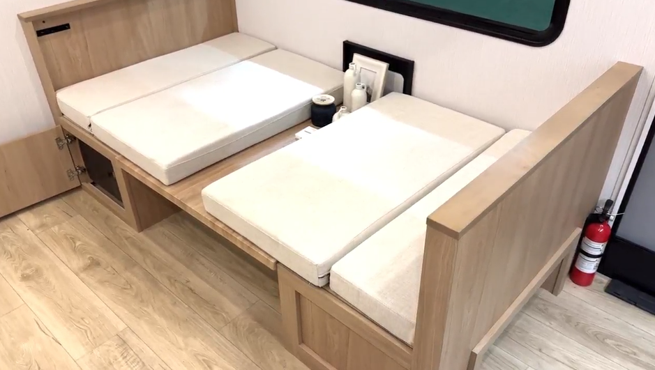 A wooden RV dinette with light beige cushions converted into a bed, with storage compartments underneath and a fire extinguisher visible nearby on the floor.