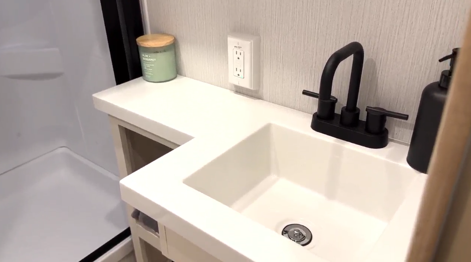 Modern RV bathroom sink with a matte black faucet, white countertop, GFCI outlet, candle, and part of a shower stall visible to the left.
