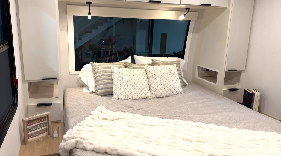 Cozy RV bedroom with a large window, neutral bedding, built-in nightstands, overhead reading lights, and decorative pillows and accessories.