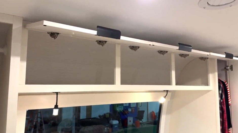 Overhead RV storage compartments above a large bedroom window, featuring multiple open cubbies with lifted cabinet doors and interior hinges.