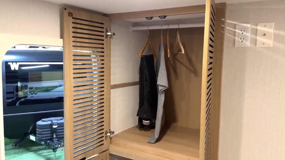  Open RV wardrobe with slatted wood doors, hanging rod, and a few clothes on wooden hangers, next to a window and power outlets.