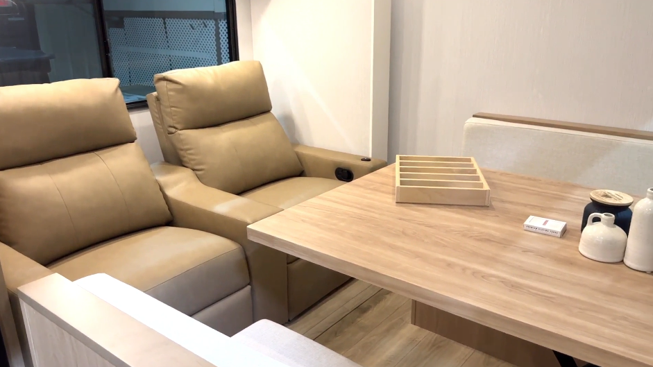 Interior of an RV slide closed and in road mode, showing a pair of tan leather recliners next to a wood dining table and bench seating, with a window in the background.
