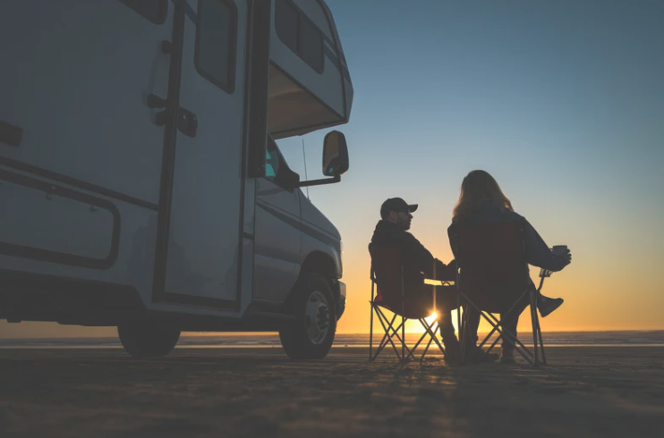 Silhouetted couple sitting in camping chairs next to their motorhome at sunset, enjoying the peaceful beach setting. The RV is parked on sand, and the horizon glows with the fading light of day.










