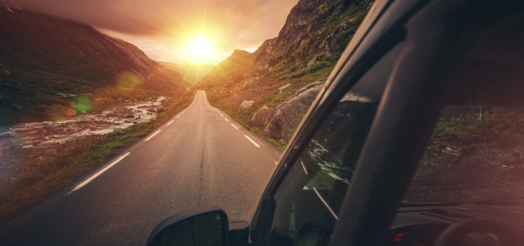 A scenic view from inside a vehicle driving down a winding mountain road at sunset. The road is flanked by rocky hills and a stream running alongside it on the left. The sun is setting in the distance, casting a warm golden glow across the valley and creating a dramatic, adventurous atmosphere.