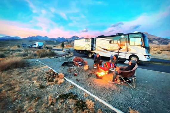 A Class A motorhome is parked in a scenic mountain campground at sunset. Three people sit around a campfire in camping chairs, enjoying the outdoors. Another RV is parked in the background, with wide open plains and mountains under a colorful sky.