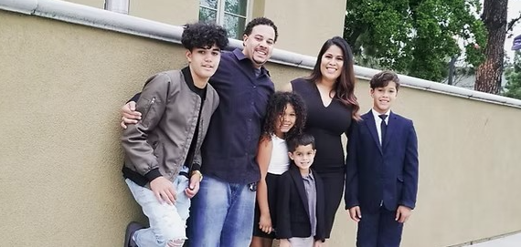 A smiling family of six stands together outside, leaning against a tan wall. The group includes two adults and four children, with everyone dressed in a mix of casual and semi-formal outfits. The parents stand in the center with their kids around them, all posing closely and happily for the photo.