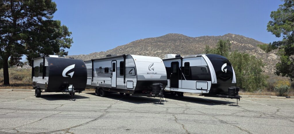 Three Wayfinder RV models are parked side by side in a lot, framed by trees and a mountainous backdrop. From left to right: a compact single-axle unit with a rounded black front, a longer mid-sized travel trailer with white siding and black accents, and a large, sleek, modern design featuring a bold black front cap and panoramic windows.