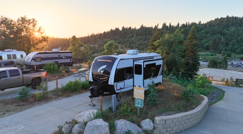 A white and black Brinkley travel trailer is parked at an elevated RV site during sunset, surrounded by greenery and trees. A gray pickup truck and additional RVs are visible nearby, with a paved driveway and a "STOP – Sites 14-57" sign in front of the trailer.