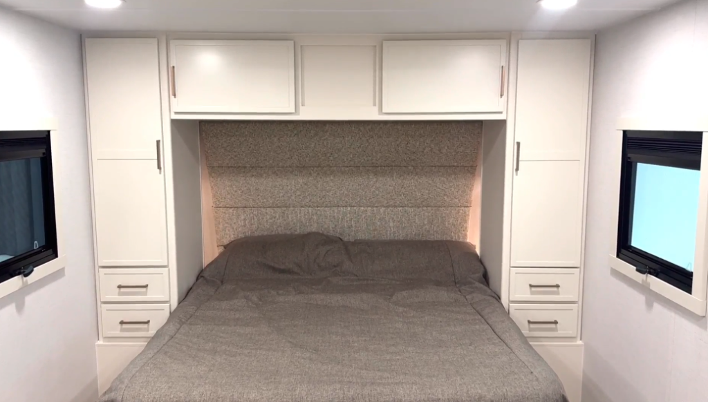 “RV bedroom with a large bed covered in a gray bedspread, surrounded by white cabinetry. Overhead storage cabinets span above the bed, while tall wardrobes with drawers are built on each side. Two windows on either side of the bed allow natural light, and recessed ceiling lights provide bright illumination. A padded, fabric-upholstered headboard adds a modern, cozy touch.