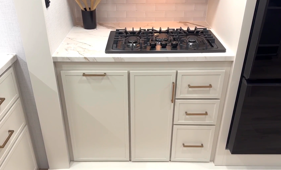 A modern RV kitchen setup featuring a sleek three-burner black stovetop with gold-trimmed control knobs, set into a white marble-look countertop. Below the counter are multiple storage cabinets and drawers with gold handles, positioned next to a glossy black refrigerator. The floor has a light wood finish, adding warmth to the clean, contemporary design.