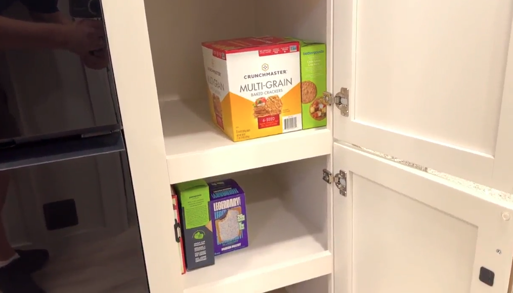 RV pantry cabinet with white doors open, showing shelves stocked with boxed snacks including Crunchmaster multi-grain crackers and other packaged items, next to a black refrigerator.