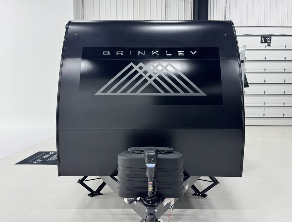 Front view of a black Brinkley travel trailer inside a showroom, featuring the Brinkley logo with geometric mountain design on the front cap, dual propane tanks with a protective cover, and an electric tongue jack at the hitch.