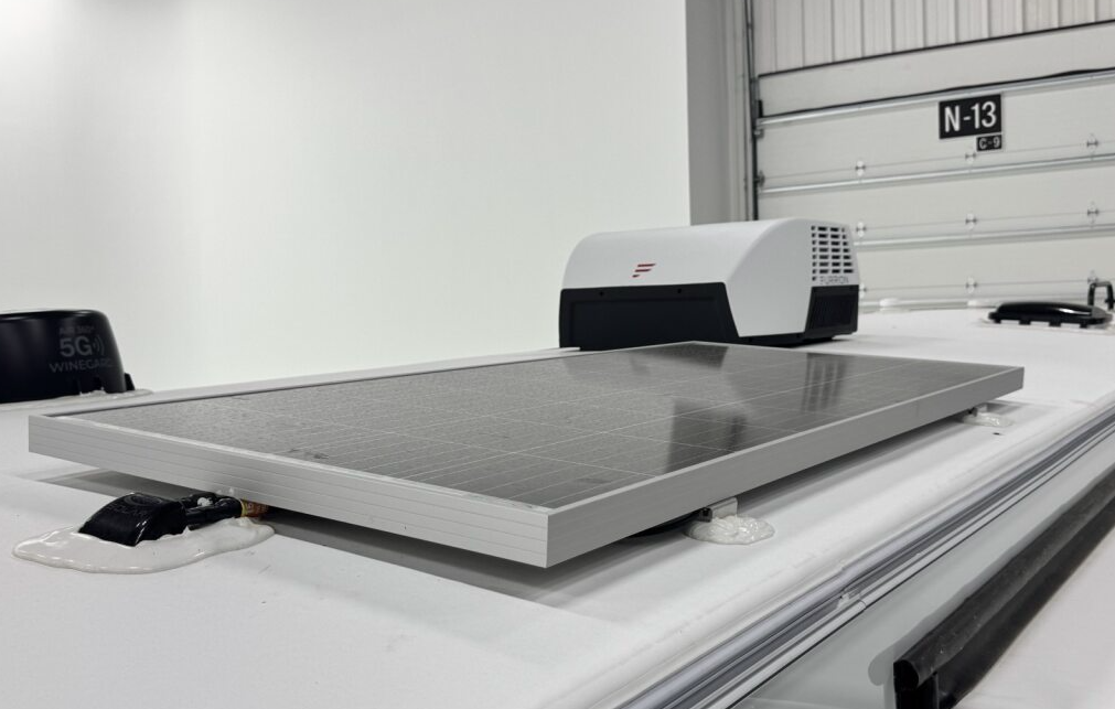 Close-up view of an RV roof featuring a mounted solar panel, with a white air conditioning unit and Winegard 5G antenna visible in the background, inside a well-lit garage.