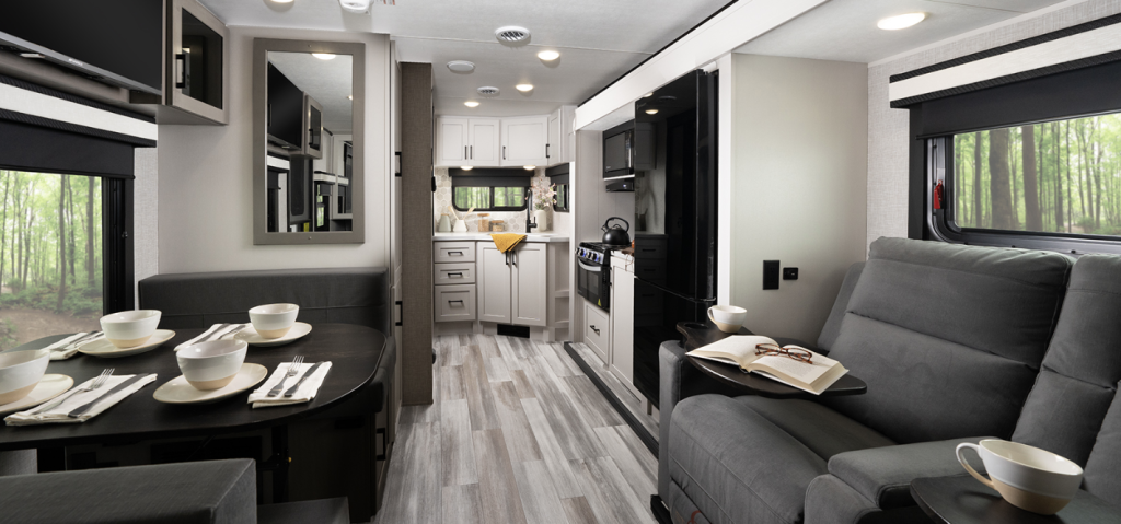 Interior of a modern RV showcasing a bright, open layout with a fully equipped kitchen, a booth-style dinette set for four with dishes and cups, and a pair of gray reclining theater seats with a book and coffee cup placed on the armrest. Large windows on both sides bring in natural light and views of a wooded forest outside.