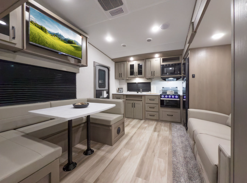 Interior of a modern RV featuring a spacious kitchen with light wood cabinetry, black hardware, a built-in microwave, oven, and stovetop. A large booth dinette with white tabletop and beige cushioned seating sits on the left under a mounted flat-screen TV. On the right, a light beige sofa faces the dining and kitchen area. The space is accented with wood-look vinyl flooring and recessed lighting, creating a clean, comfortable, and functional living space.