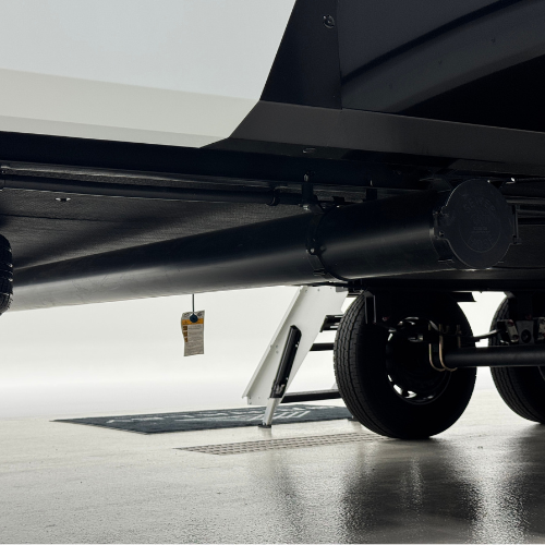 Undercarriage view of a Brinkley RV showing the enclosed underbelly with a visible cylindrical waste pipe system, sturdy suspension, dual axles, and black tires. A portion of the entry steps can also be seen extending to the floor.