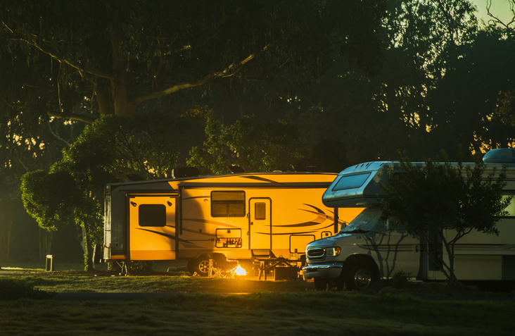 Two RVs are parked at a wooded campsite during dusk, with one brightly lit travel trailer glowing yellow from interior lights and a campfire burning warmly in front. A motorhome sits nearby under the trees, surrounded by a peaceful outdoor setting.