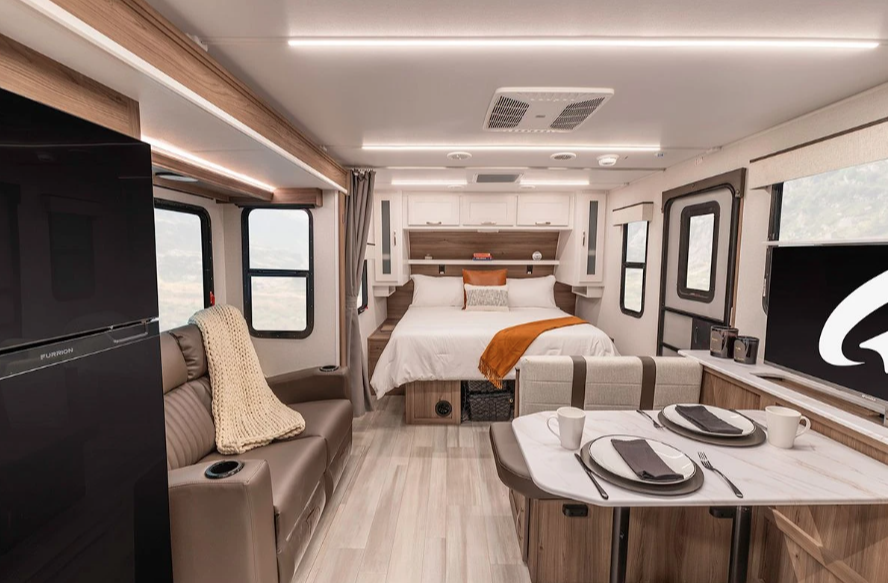 Interior of a modern travel trailer featuring a cozy queen bed with accent pillows, a dining booth set for two, and a reclining sofa with a throw blanket. Large windows bring in natural light, highlighting the stylish wood finishes and compact RV design.