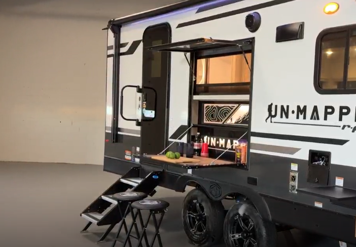 A modern travel trailer with dual axles is shown indoors, featuring a fold-out outdoor kitchen setup. The side panel is lifted to reveal a counter with condiments, limes, and a drink bottle, while two stools are placed in front of the counter. The trailer has bold black wheels, sleek white and black exterior graphics, and the brand name "UN•MAPPED" displayed prominently.