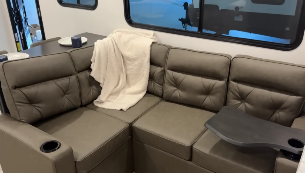 Modern RV interior featuring a dark gray sectional sofa with tufted cushions. A cream-colored throw blanket is draped over the backrest, and a small detachable table with a cupholder is attached to one seat. A large window lets in natural light, with a dining area partially visible in the background, creating a cozy and functional living space.