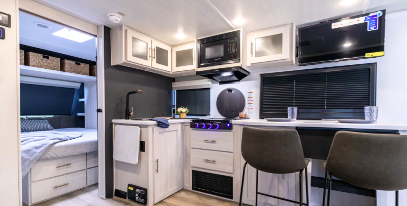 Interior of R-Pod Unmapped travel trailer showing kitchen with microwave, stove, bar seating, and view into bedroom with Olympic queen bed and overhead storage.