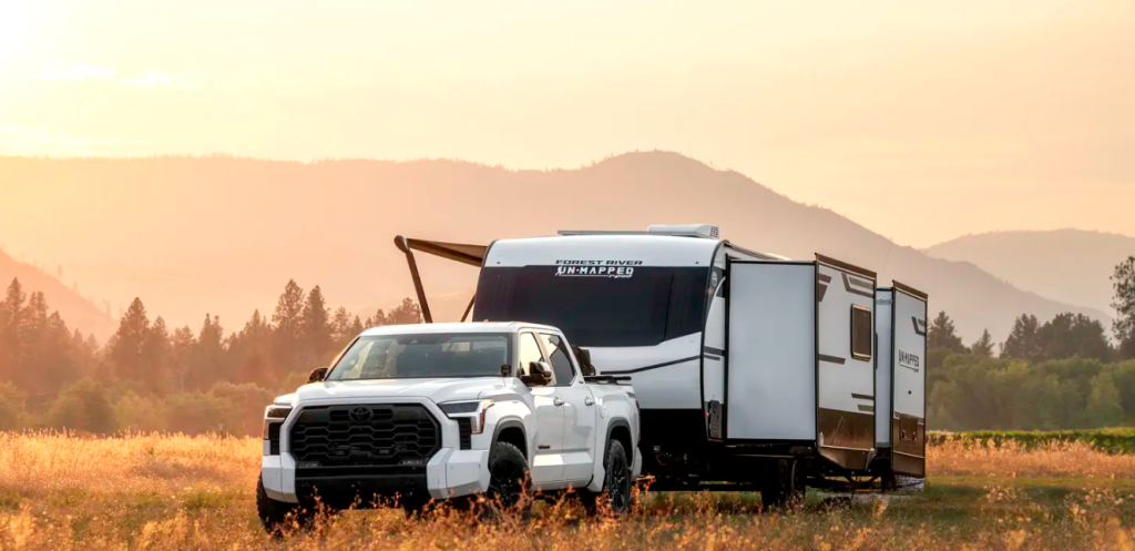 Forest River R-Pod Unmapped travel trailer with slide-outs, towed by a white truck across a scenic mountain field at sunset.