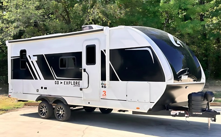 "White Wayfinder Go Explore Limited travel trailer with sleek black accents and large panoramic front window, parked on pavement with green trees in the background.