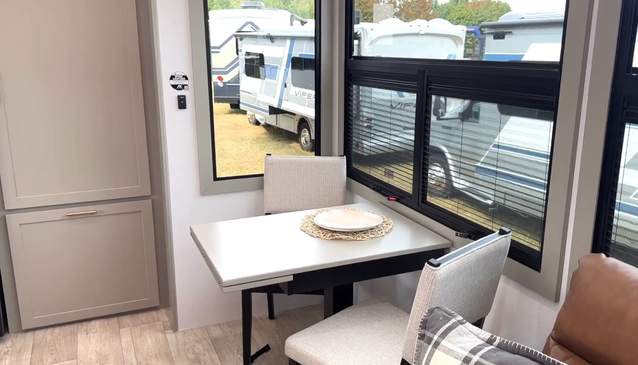 Cozy RV dining nook in the Axiom Imperium 12900 featuring a two-seat table by large panoramic windows with natural light and modern cabinetry.
