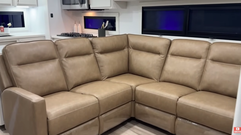 Brinkley RV living room with large tan leather sectional sofa, modern kitchen in background, and panoramic windows with blackout shades.