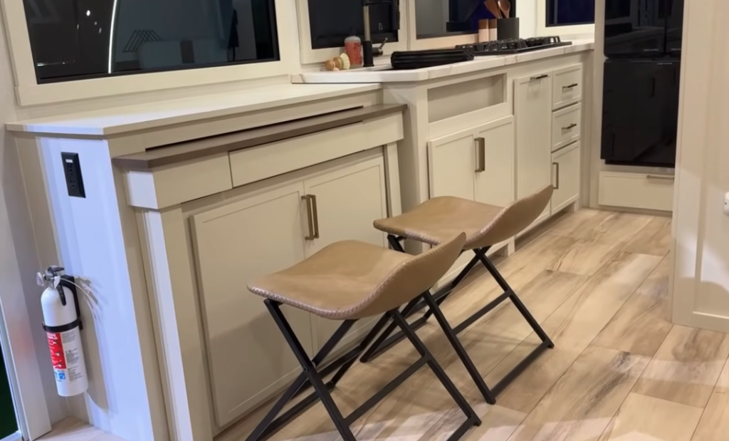 Brinkley RV kitchen with light cabinetry, built-in counter storage, modern cooktop, and two leather barstools for casual dining.