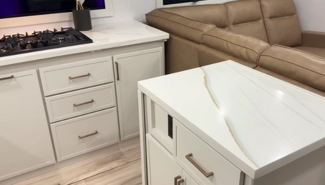 rinkley RV kitchen interior with white cabinetry, solid-surface countertops, built-in gas cooktop, and adjacent leather theater seating.