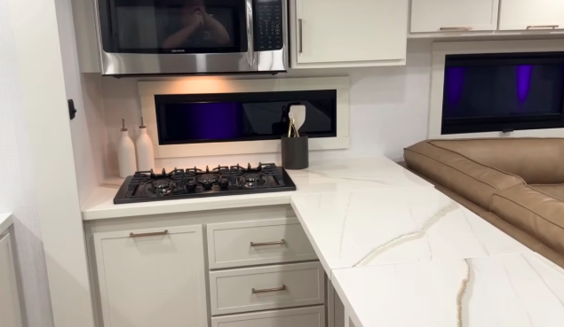 Brinkley RV kitchen with solid-surface countertops, built-in gas cooktop, overhead microwave, and modern white cabinetry beside leather seating.