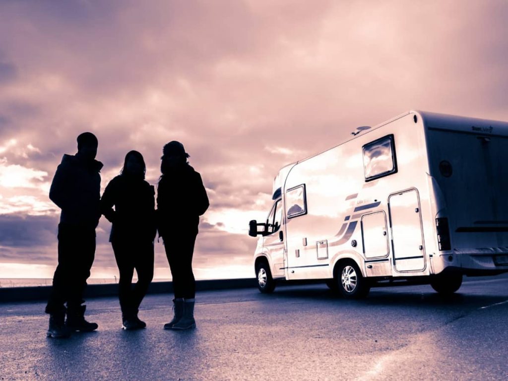 Three people stand together near a motorhome parked on an open road at sunset, their silhouettes outlined against a dramatic purple-toned sky.