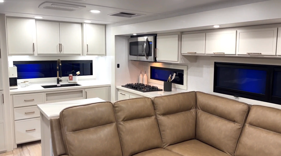 Modern RV kitchen inside the Brinkley Model I 280 featuring solid-surface countertops, white cabinetry with brushed metal hardware, a 12V refrigerator, and a built-in 3-burner cooktop with convection microwave. A tan sectional sofa sits in the foreground, showing the open-concept design that connects the kitchen and living area. Large horizontal windows and LED lighting create a bright, residential feel.