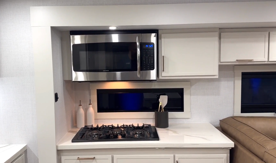 Kitchen cooking area inside the Brinkley Model I 280 featuring a stainless steel convection microwave above a three-burner gas cooktop with solid-surface counters and white cabinetry. Soft lighting under the overhead cabinet and a narrow horizontal window create a bright, modern, and functional workspace ideal for cooking on the road.